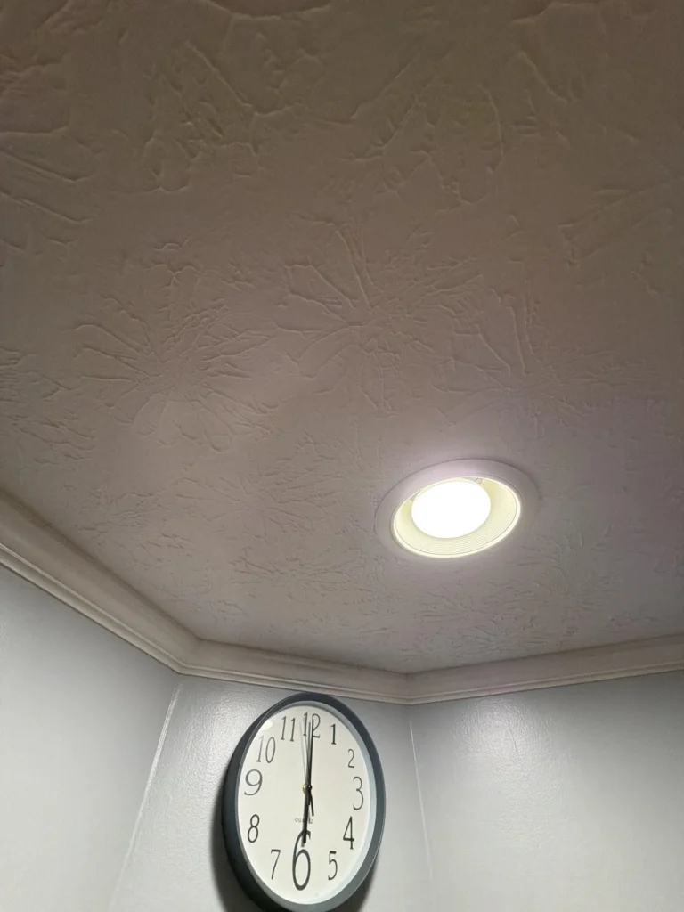 Looking up at hallway ceiling showing skip-trowel texture with visible water stain and discoloration around recessed light fixture, white crown molding, wall clock below showing time