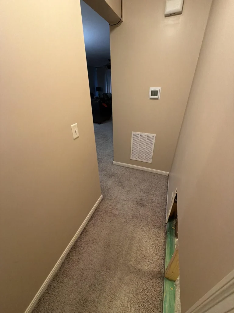 Narrow carpeted hallway with beige walls, HVAC return air vent and thermostat panel on end wall, drywall cutout at baseboard on right side showing wood framing and subfloor during water damage repair
