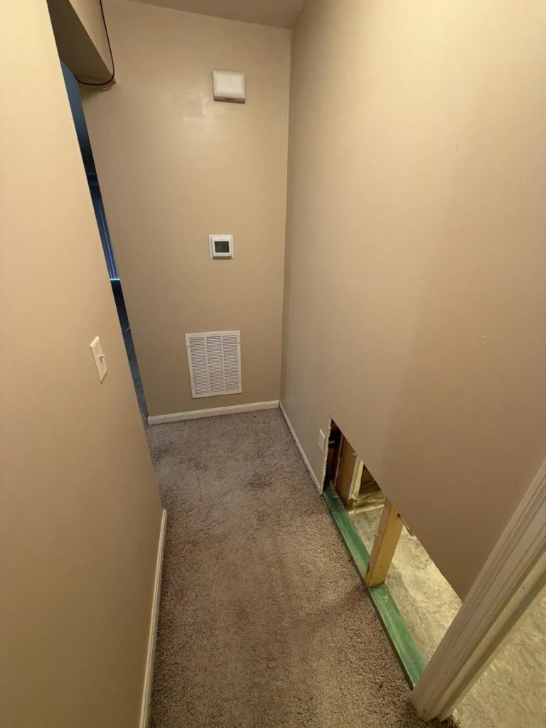 End of carpeted hallway with beige walls showing HVAC return air vent and thermostat on far wall, drywall cutout at baseboard level on right exposing wood framing, light switch on left wall