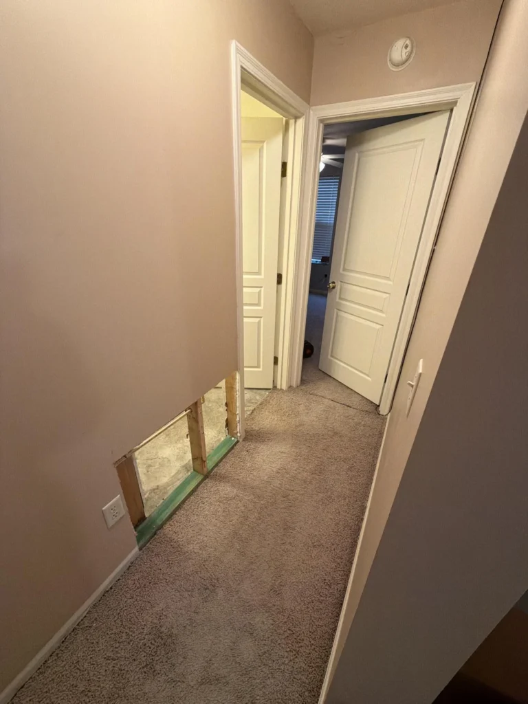Carpeted hallway with beige walls showing two open interior doors leading to rooms, drywall cutout at baseboard level on left side exposing wood framing and concrete subfloor
