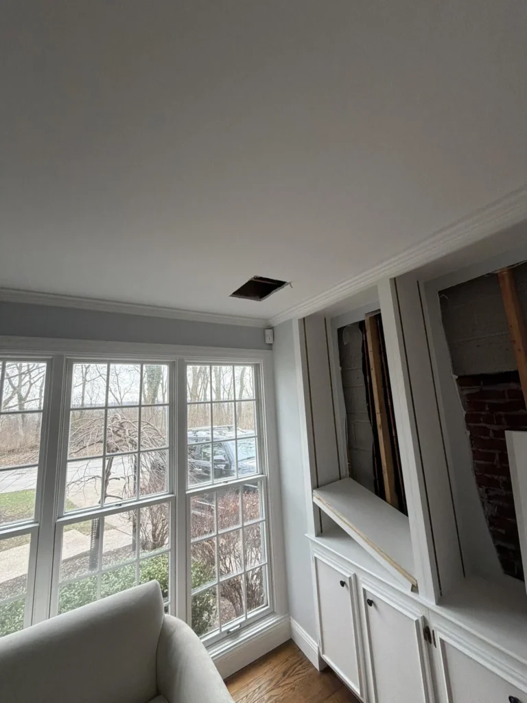 Home office corner view showing a ceiling access hole from water damage, white built-in cabinets with upper portion open revealing an exposed cinder block wall and wooden studs, large multi-pane windows overlooking a snowy yard, and a cream armchair in the foreground