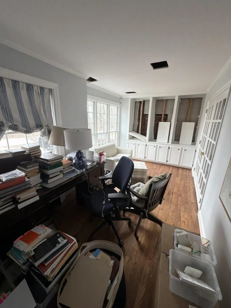 Wide view of a home office with two ceiling access holes from water damage, a white sofa with a tufted ottoman in front of large multi-pane windows, a cluttered dark wood desk with books and lamps, an office chair, built-in cabinets with exposed wall cavities, and hardwood floors
