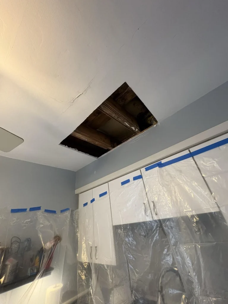 Kitchen ceiling with a large rectangular drywall cutout exposing dark water-stained wooden joists and subfloor above, white upper cabinets covered in plastic sheeting below, ceiling cracks visible