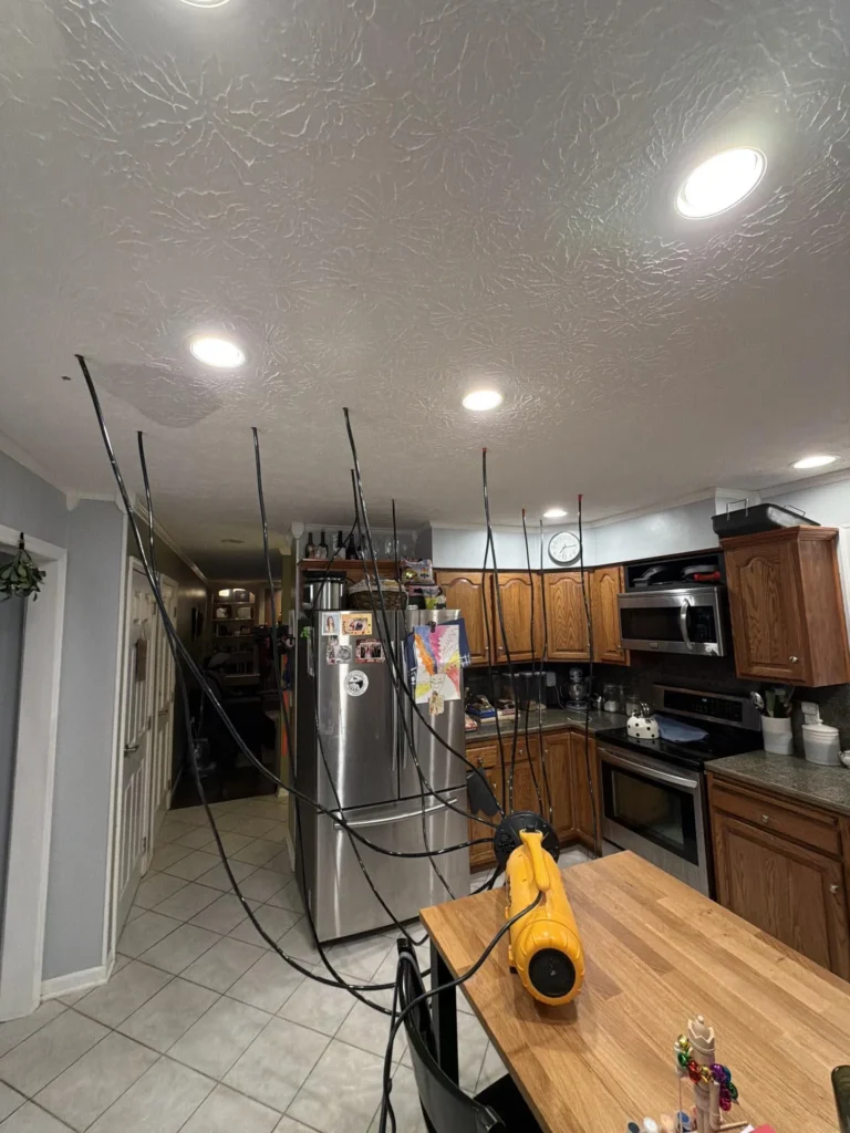 Wide kitchen view showing water damage drying in progress with yellow PDS-12 blower on island, multiple black drying hoses inserted into ceiling at injection points throughout skip-trowel ceiling, stainless appliances and oak cabinets visible