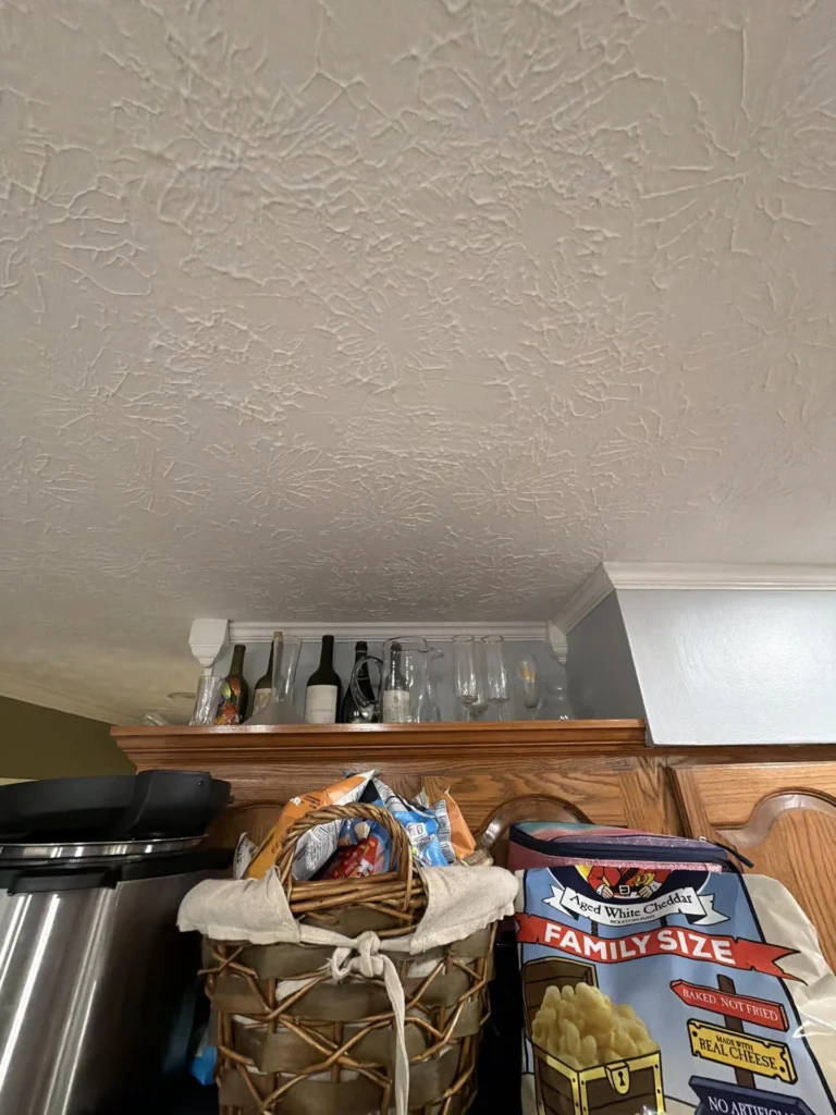 Kitchen ceiling showing skip-trowel texture above oak kitchen cabinets, elevated wine bottle and glass rack shelf with bottles and stemware mounted near crown molding, wicker basket and snack items on top of refrigerator below