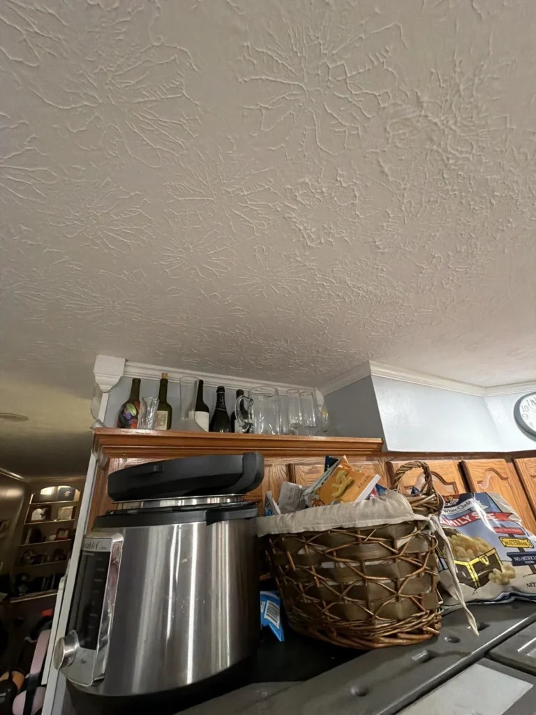 Kitchen ceiling with skip-trowel texture above stainless French door refrigerator and oak upper cabinets, elevated wine rack shelf with bottles near crown molding, stainless appliances, wicker basket and food items on refrigerator top
