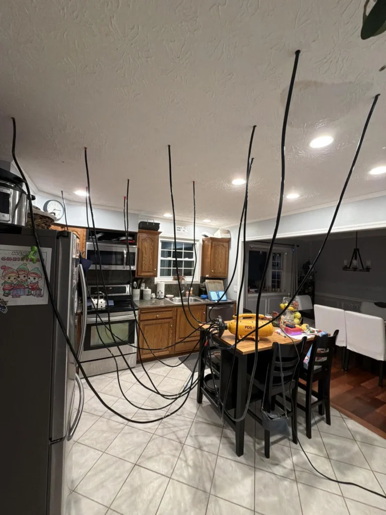 Full view of kitchen and adjacent dining room during water damage remediation showing yellow PDS-12 drying blower on island with numerous black hoses injected into skip-trowel ceiling across entire span, refrigerator on left, white dining chairs in background