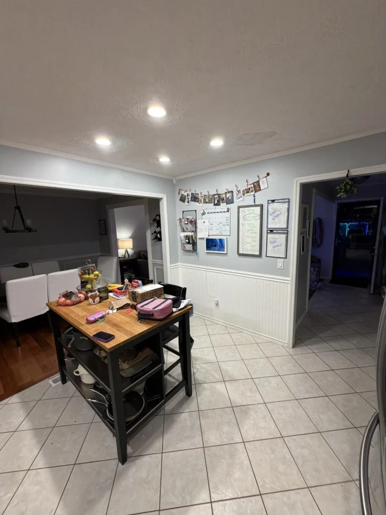 Kitchen eating area at night with light blue walls, white wainscoting, crown molding, recessed lighting, butcher block island with bar stools, family organization wall with photo garland and whiteboards, doorway to dark hallway, and tile floor