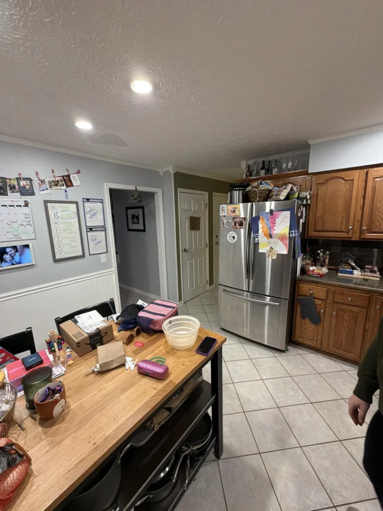 Kitchen with butcher block island cluttered with items, stainless French door refrigerator covered with family photos and magnets, oak upper and lower cabinets with granite counters, recessed lighting, open doorway to hallway, tile floor
