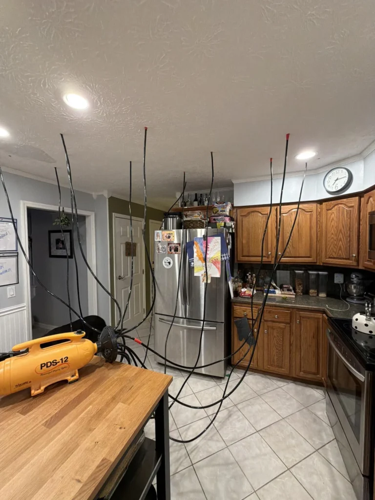 Kitchen during water damage drying showing yellow PDS-12 desiccant drying system on butcher block island with multiple black flexible hoses running up to injection points in skip-trowel ceiling, stainless refrigerator and oak cabinets in background