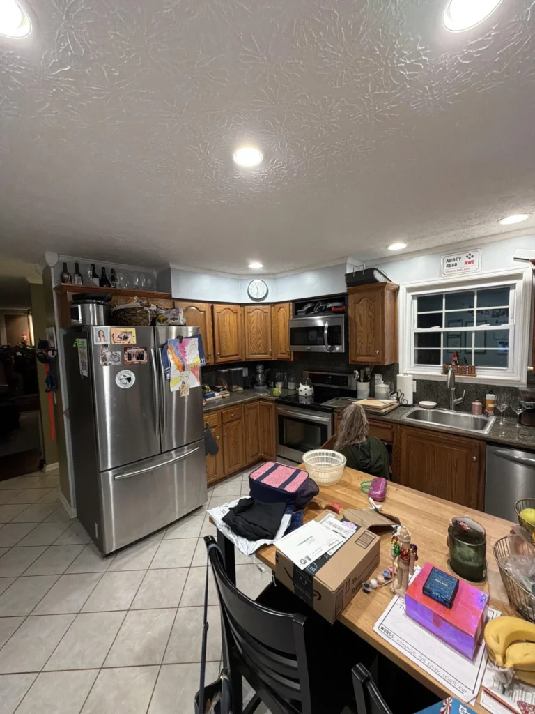 Wide kitchen view showing full L-shaped layout with stainless French door refrigerator, oak raised panel cabinets, stainless range and microwave, dark granite countertops, recessed lighting, single window over sink, tile floor, butcher block island in foreground