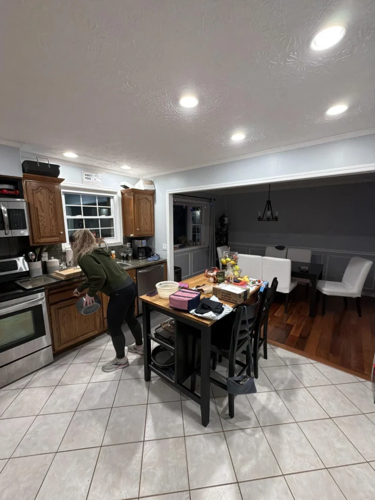 Kitchen with person at sink wearing green hoodie, stainless appliances, oak cabinets, tile floor, butcher block island with bar chairs, open pass-through to dining room with white upholstered chairs and dark walls, recessed lighting throughout