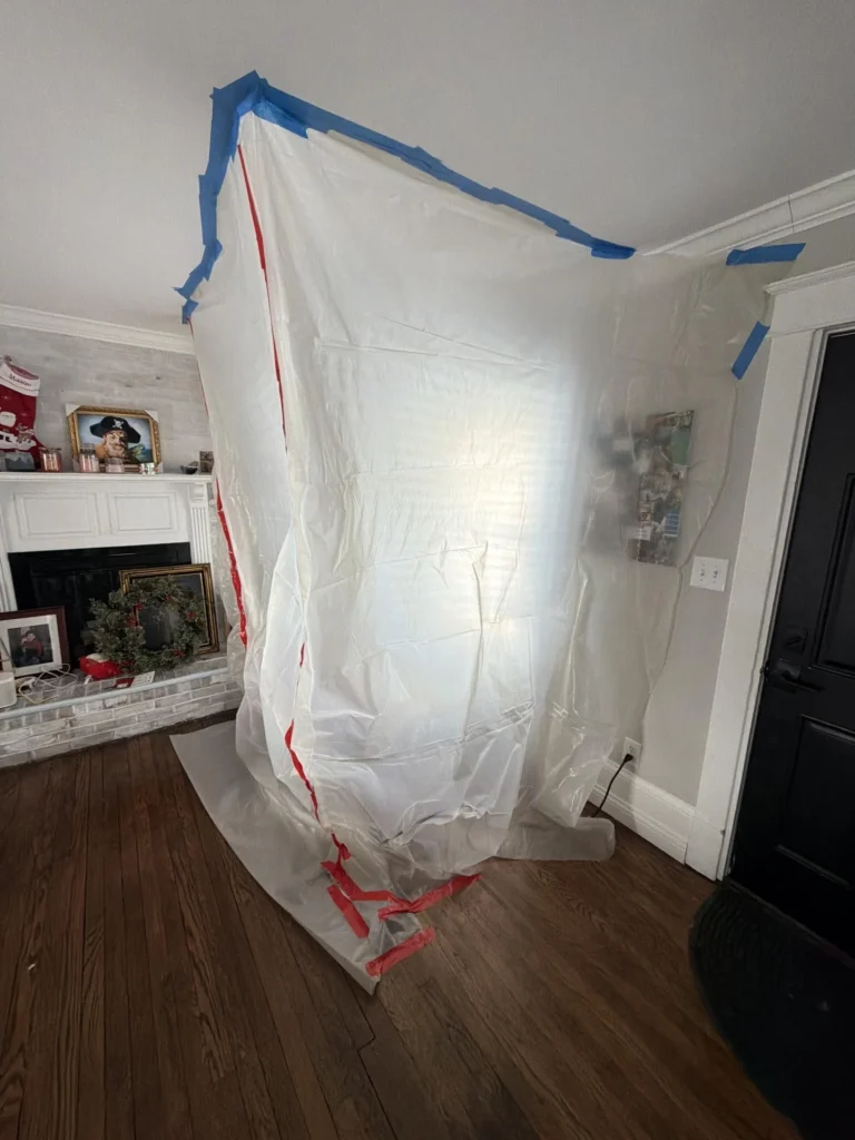 Living room with a large white plastic containment barrier taped to the ceiling and wall with blue painter's tape and red zip ties at the base, white brick fireplace with holiday wreath and decorations visible to the left, hardwood floor