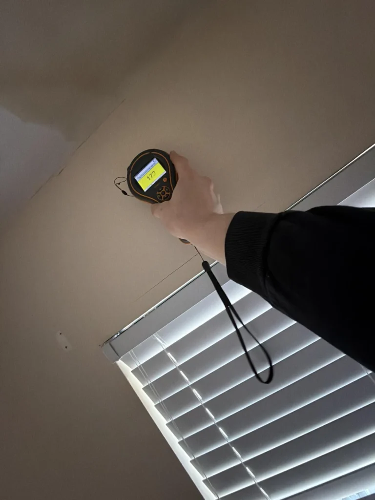 Hand holding moisture meter showing yellow 172 reading against ceiling drywall near window with horizontal blinds, indicating elevated moisture content