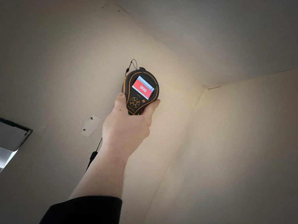 Hand holding pinless moisture meter showing maxed-out red 999 reading pressed against water-damaged drywall ceiling corner, indicating severe saturation