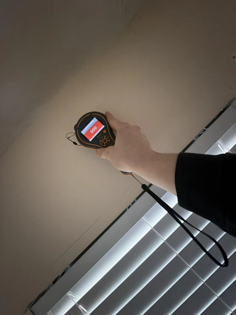 Hand holding moisture meter displaying maxed red 999 reading against ceiling-wall junction above window with horizontal blinds, confirming severe water intrusion