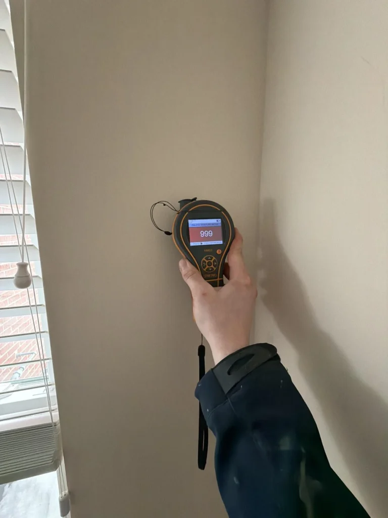 Hand holding pinless moisture meter showing maxed red 999 reading on interior drywall wall near window with blinds, confirming extreme water saturation