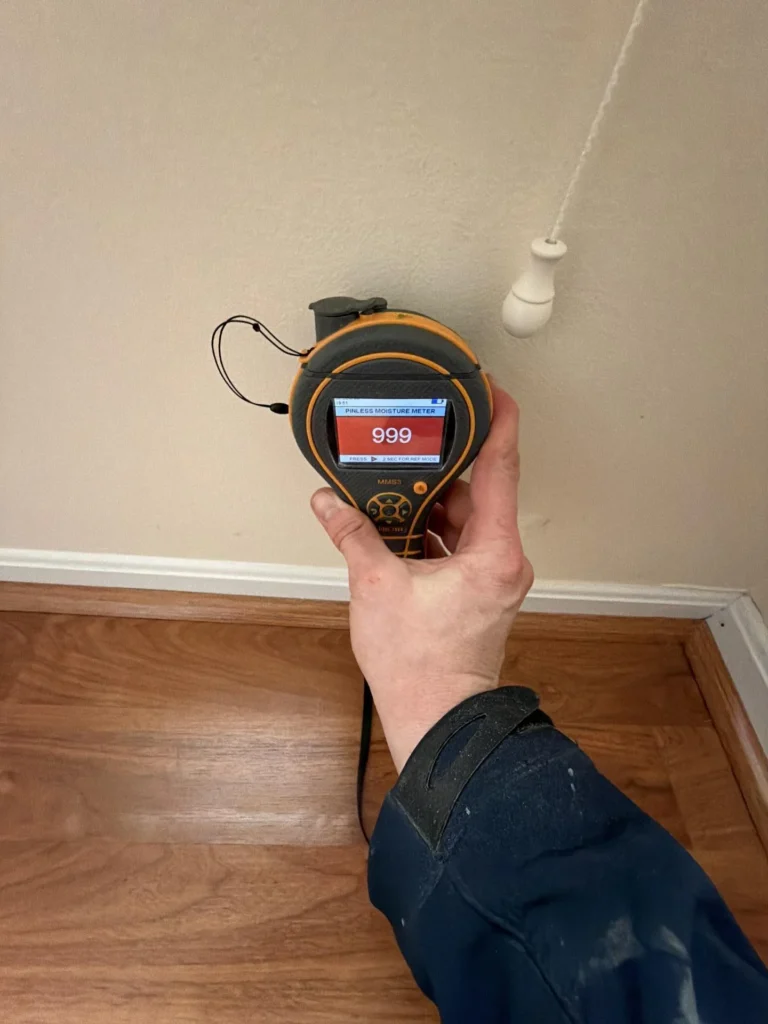 Hand holding moisture meter displaying maxed red 999 reading against lower wall drywall near baseboard and hardwood floor, indicating severe moisture in wall cavity
