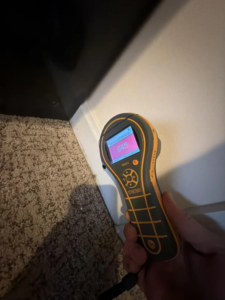 Hand holding a Protimeter MMS3 pinless moisture meter displaying a reading of 945 on a pink-highlighted screen, pressed against a white baseboard at the wall-carpet junction, indicating severely elevated moisture levels