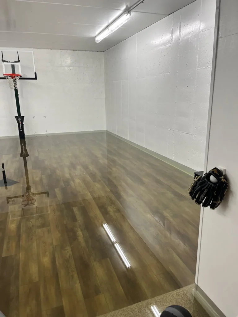 Corner view of indoor sport court with water-damaged wood-look vinyl flooring, basketball hoop visible at left, baseball glove hanging on wall hook, fluorescent strip lights reflecting in floodwater, white cinder block walls, carpet entry area at bottom