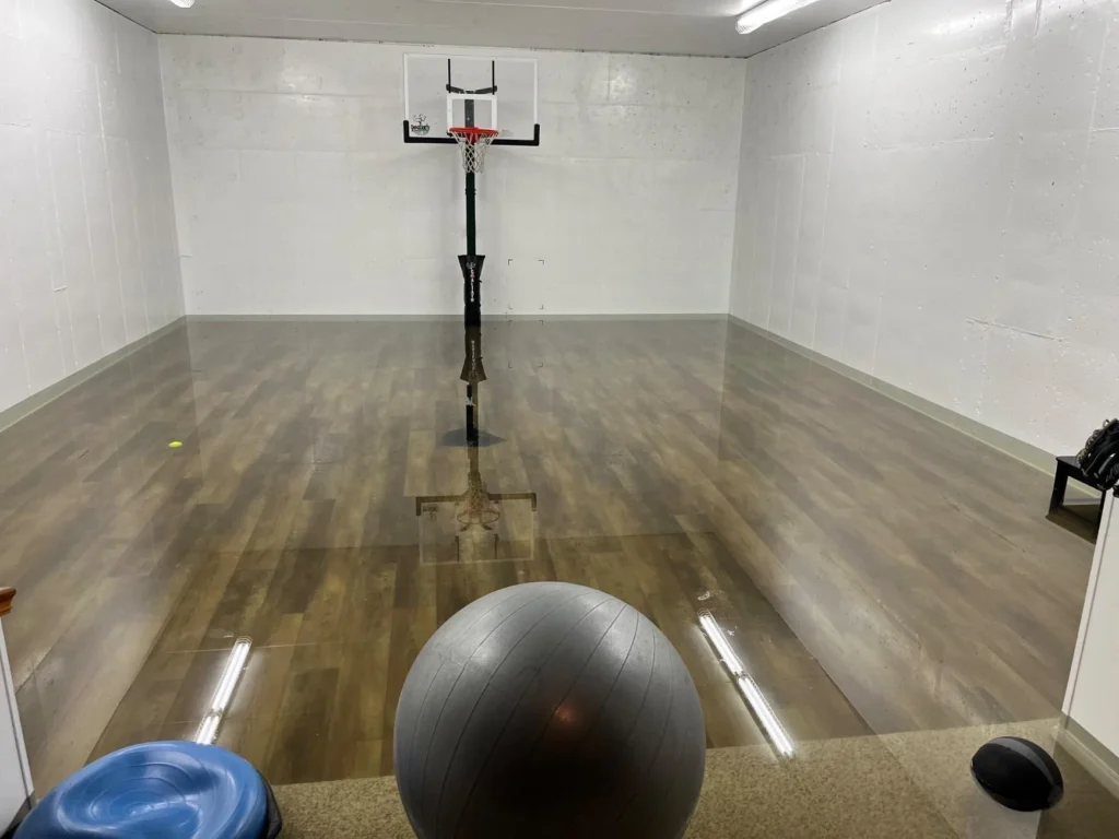 Indoor home sport court with wood-look vinyl flooring covered in standing water from flooding, portable basketball hoop with acrylic backboard reflected in floodwater, gray exercise ball and blue wobble cushion in foreground, white painted cinder block walls with fluorescent lighting