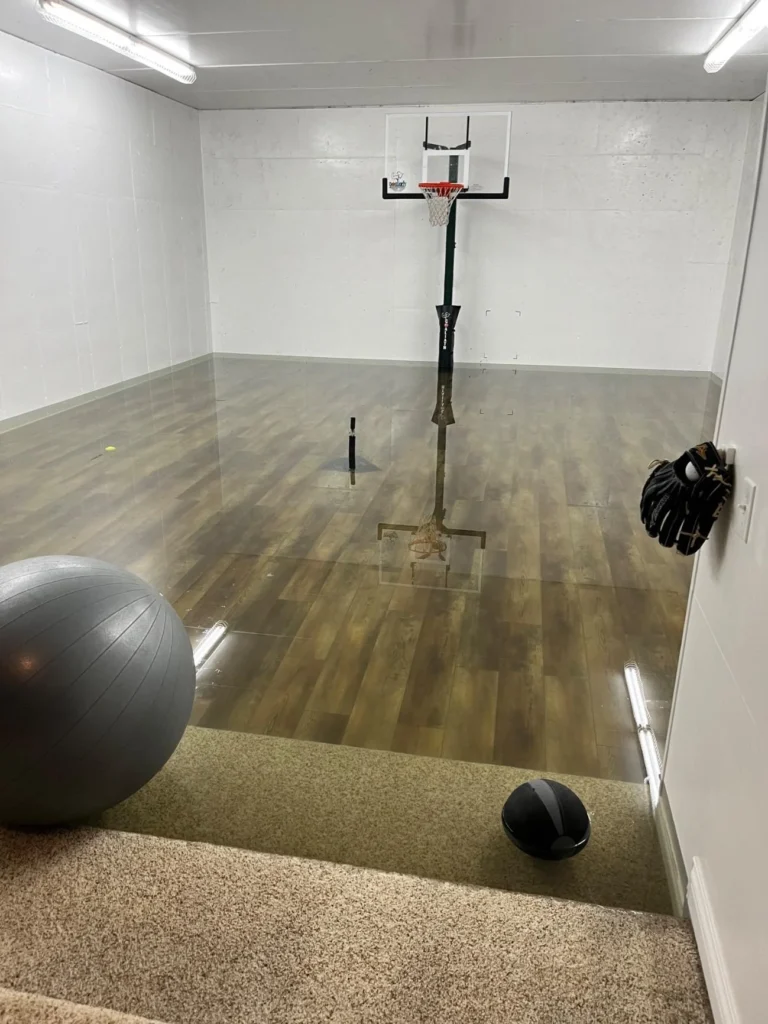 Side view of flooded indoor home sport court showing standing water on wood-look vinyl floor, basketball hoop reflected in water, gray exercise ball on carpet threshold, black medicine ball and baseball glove on wall hook, fluorescent ceiling lights
