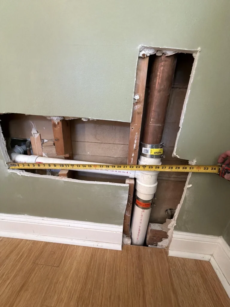 Contractor measuring the horizontal width of PVC supply and drain pipes exposed inside a green-painted wall opened for plumbing access during a bathroom renovation