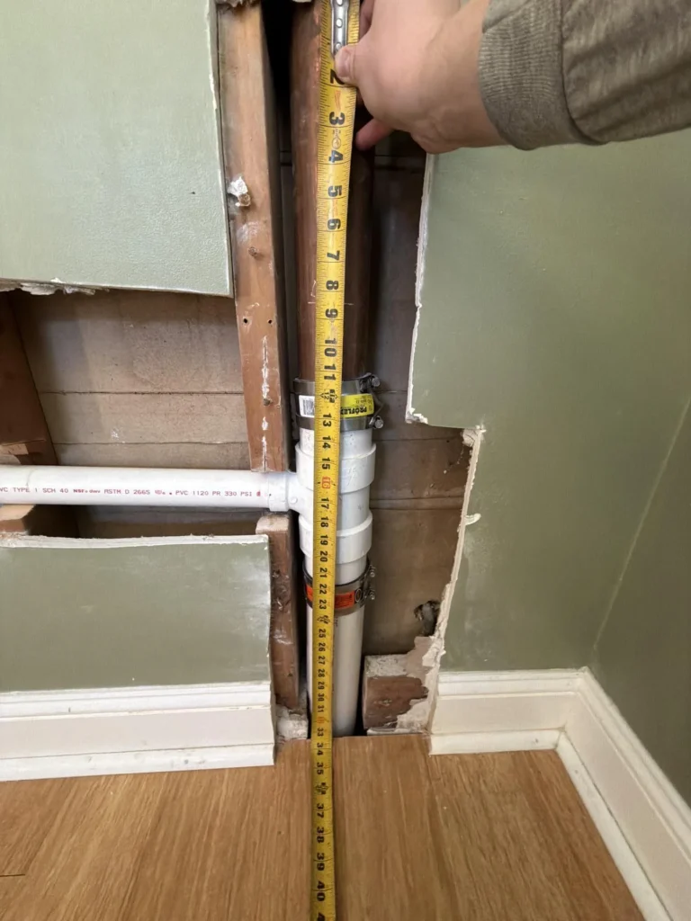 Contractor measuring the vertical height of PVC drain pipes and copper stack exposed through an opened green drywall during a bathroom plumbing repair or renovation