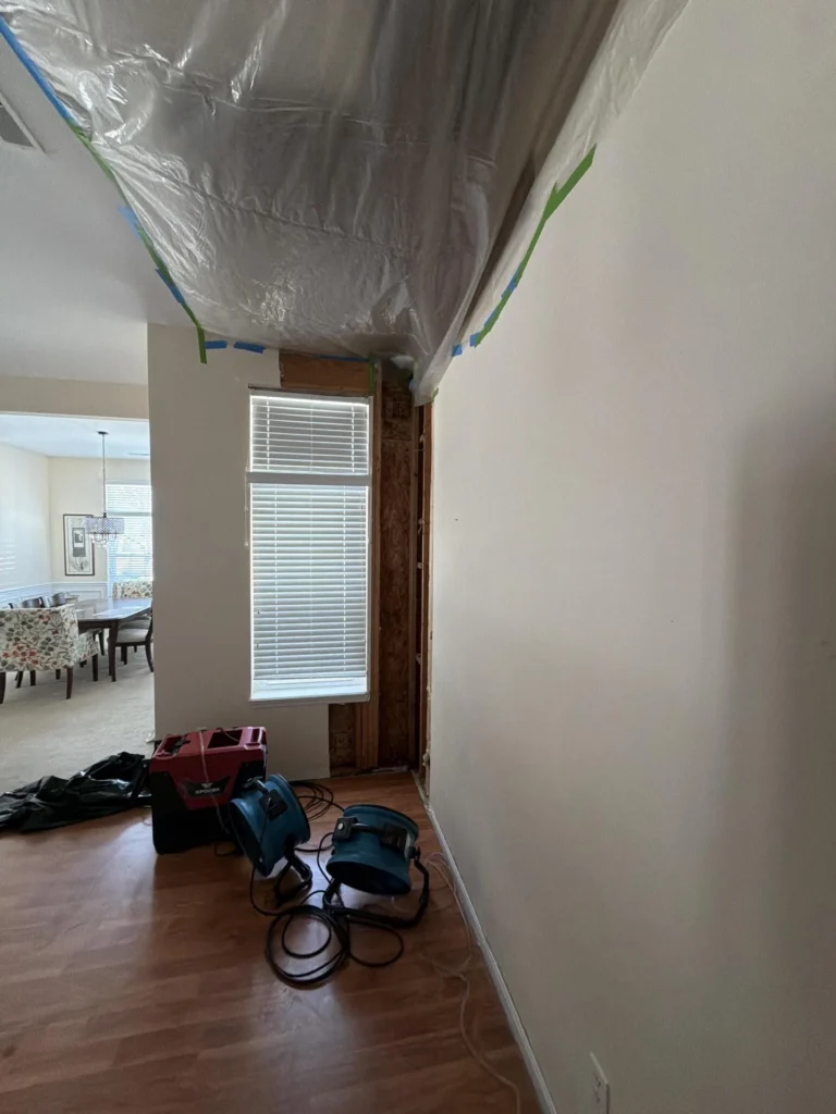 Water damage remediation in progress with plastic sheeting taped to ceiling with blue and green tape, stripped drywall revealing framing near tall window, air movers and dehumidifier on hardwood floor