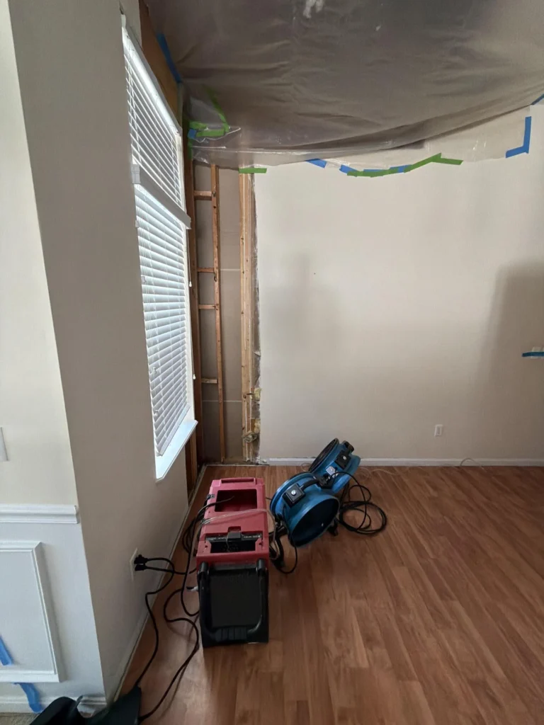 Wide view of water damage remediation in progress, stripped wall cavity beside window with plastic containment ceiling taped with blue and green tape, red dehumidifier and blue air mover on wood floor