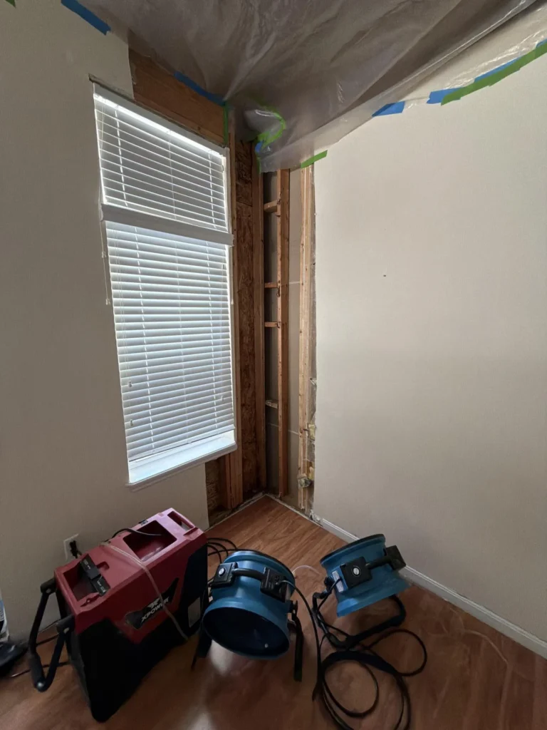 Close-up of water damage remediation showing stripped wall section with exposed wood framing and OSB beside window, plastic containment on ceiling, air movers on hardwood floor