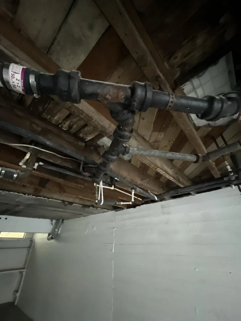 Basement ceiling showing a corroded cast-iron pipe T-junction with a rubber coupling, connected to multiple drain pipes amid severely rotted wooden floor joists and PVC supply pipes below