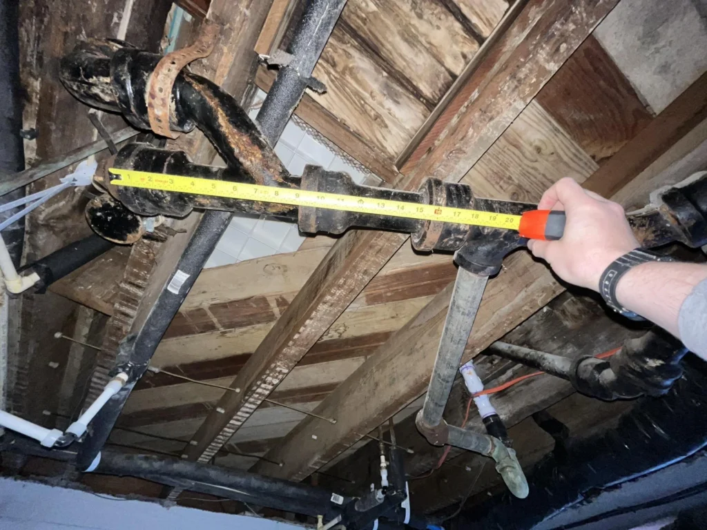 A person holds a measuring tape across a section of corroded cast-iron drain pipe in a basement ceiling, with the tape reading approximately 20 inches, surrounded by rotted joists and other pipes
