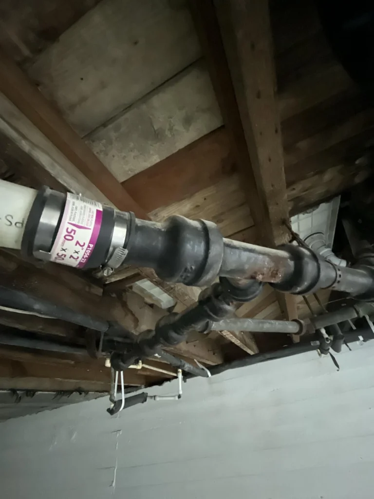 Basement ceiling view showing a complex network of old corroded cast-iron drain pipes with multiple fittings and junctions running through severely rotted wooden floor joists with PVC supply pipes below