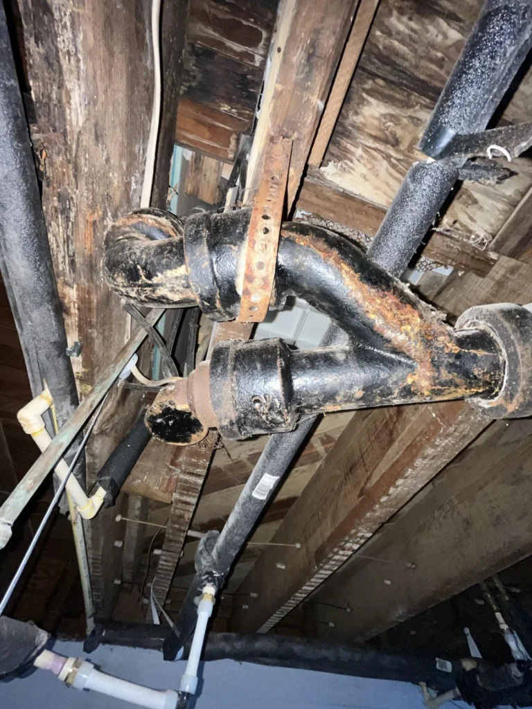 Basement ceiling view showing heavily corroded and rusting cast-iron drain pipe junction surrounded by severely rotted and water-damaged wooden floor joists, with white PVC supply pipes visible below