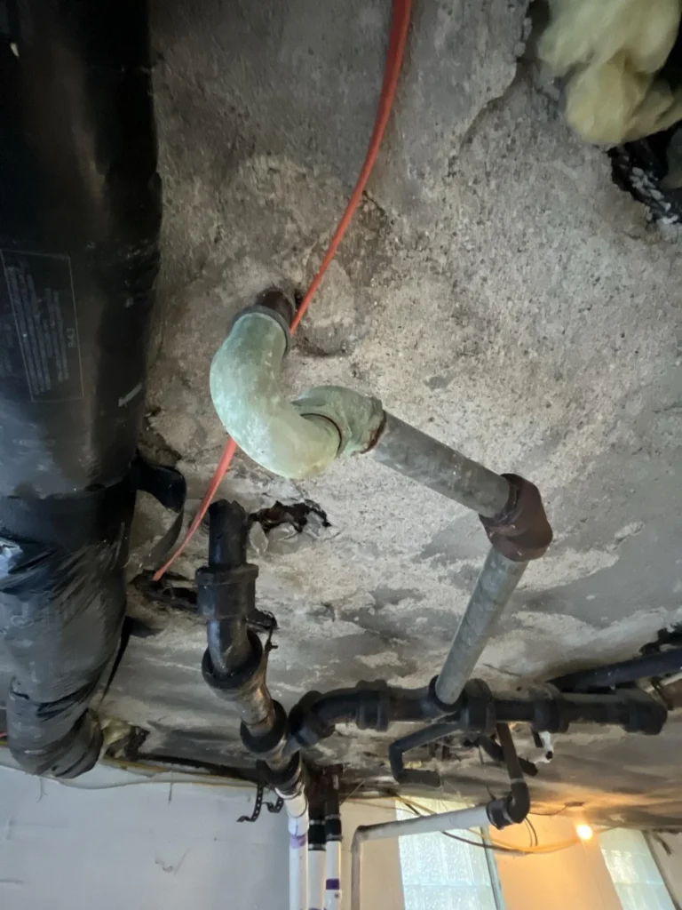 Basement ceiling view showing a heavily corroded cast-iron pipe elbow fitting with orange rust staining passing through a deteriorated concrete ceiling, with an orange wire and additional black iron pipes nearby