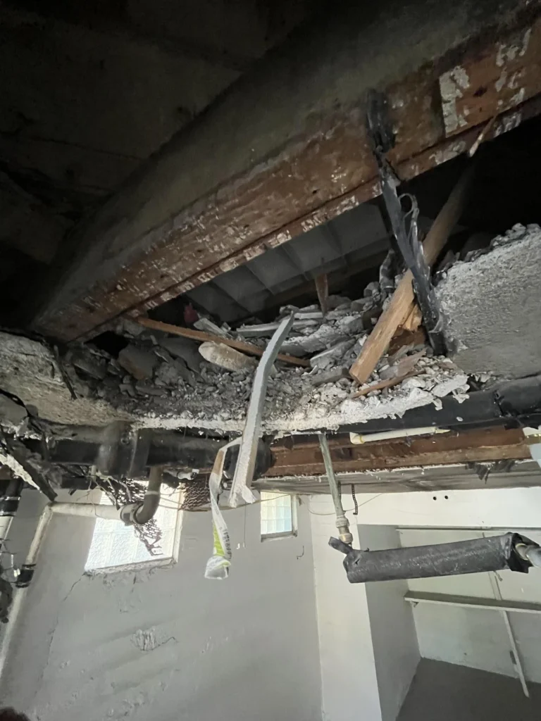 Close-up of a collapsed basement ceiling showing severely rotted wooden floor joists, large chunks of fallen plaster and drywall debris, and black drain pipes amid the destruction