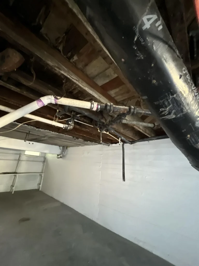 Severely deteriorated basement ceiling with large irregular holes exposing rotted wood above, alongside black drain pipes, an orange wire, and exposed insulation indicating major structural water damage