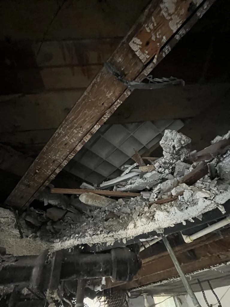 Close-up of a collapsed basement ceiling area showing severely rotted wooden floor joists with large chunks of fallen plaster and concrete debris piled between them, alongside black drain pipes and strapping hardware