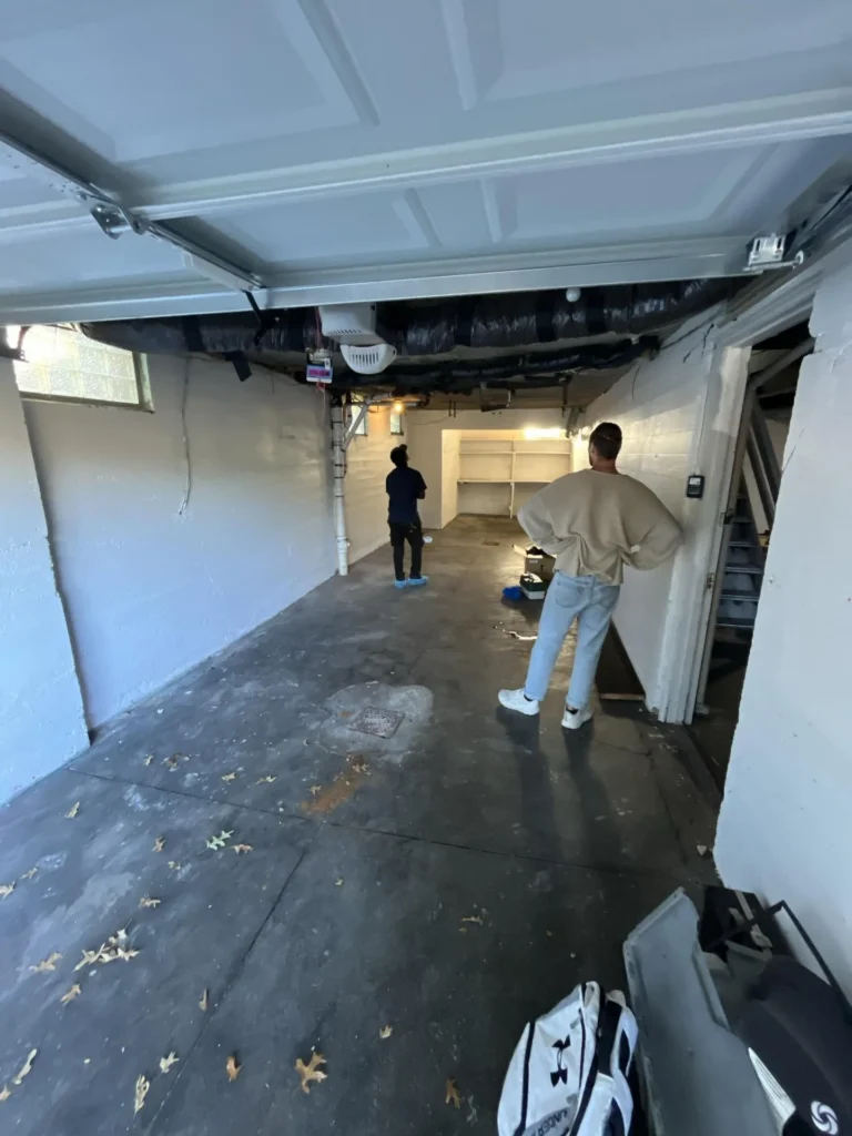 Wide view of basement ceiling showing an extensive network of corroded black cast-iron drain pipes, white PVC pipes, and an orange wire running through severely rotted wooden floor joists
