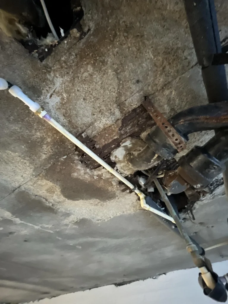 Close-up of basement ceiling showing a white PVC pipe running alongside severely corroded cast-iron pipe fittings embedded in a deteriorated concrete ceiling with rotted wood visible in the opening