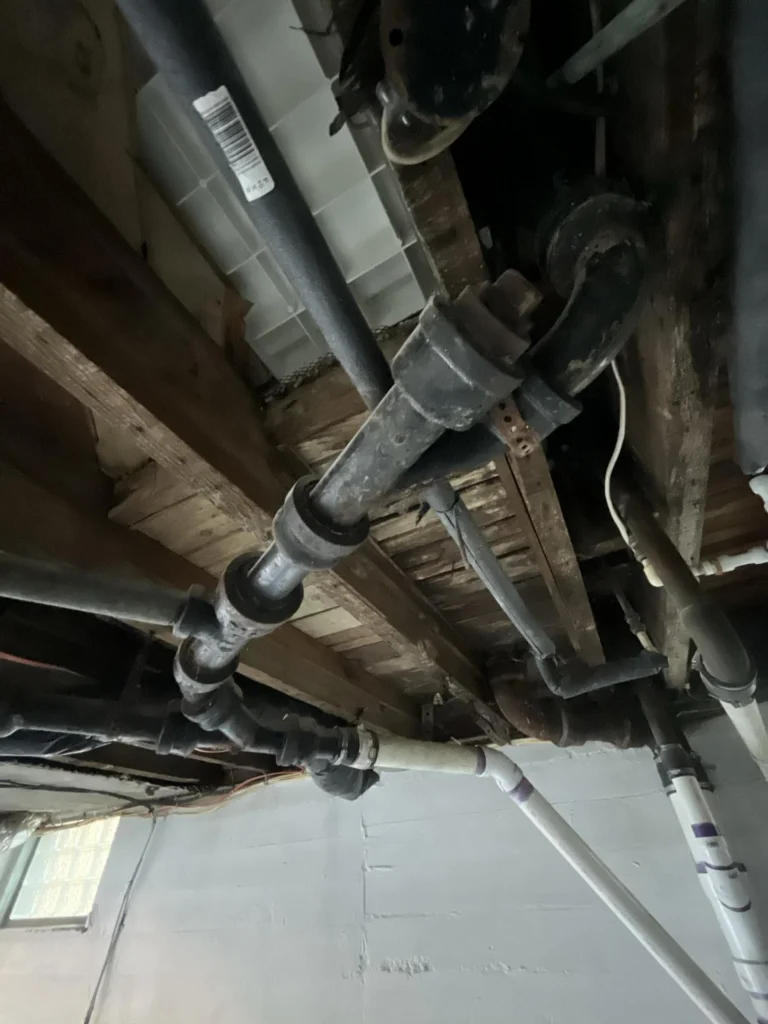 Basement ceiling with multiple holes and damaged plaster exposing the structure above, with an extensive network of black iron and PVC drain pipes alongside an orange wire running through the space