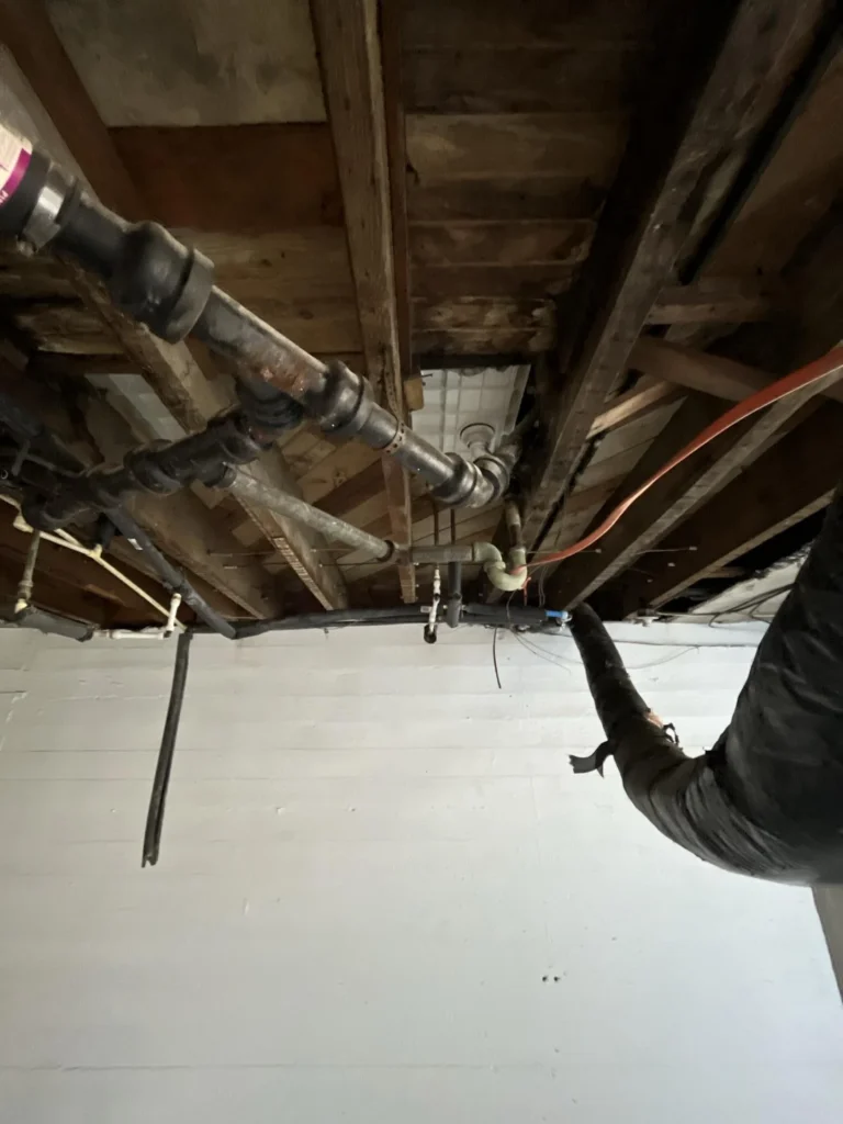 Basement ceiling corner showing a large black flexible duct penetrating through the concrete ceiling alongside an orange electrical wire and yellow fiberglass insulation, with PVC and black iron pipes below