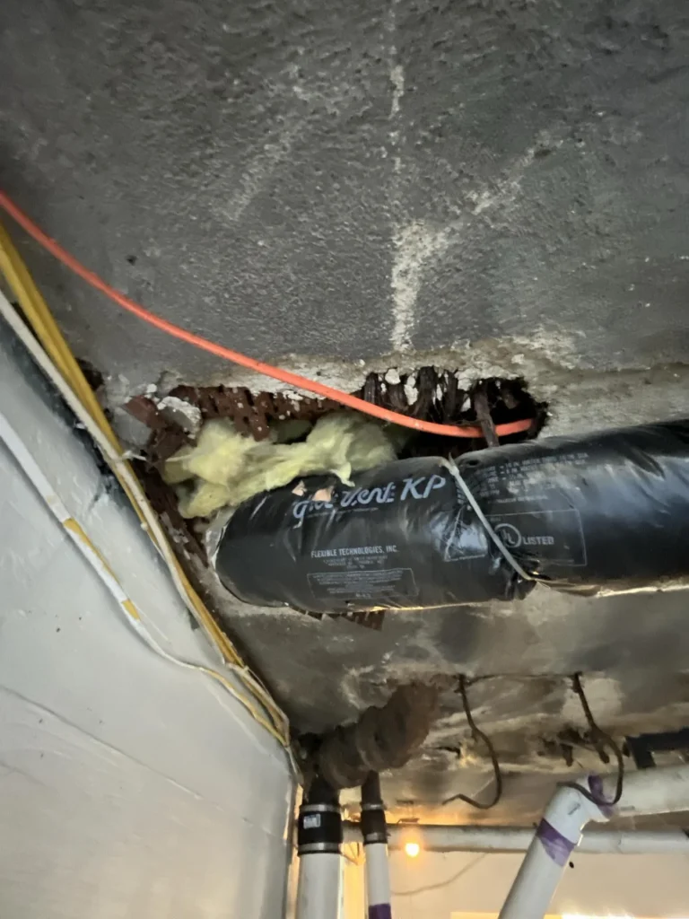 Basement ceiling showing a junction of white PVC pipe connecting to corroded cast-iron drain pipes amid severely rotted wood floor joists with rust staining and deterioration throughout