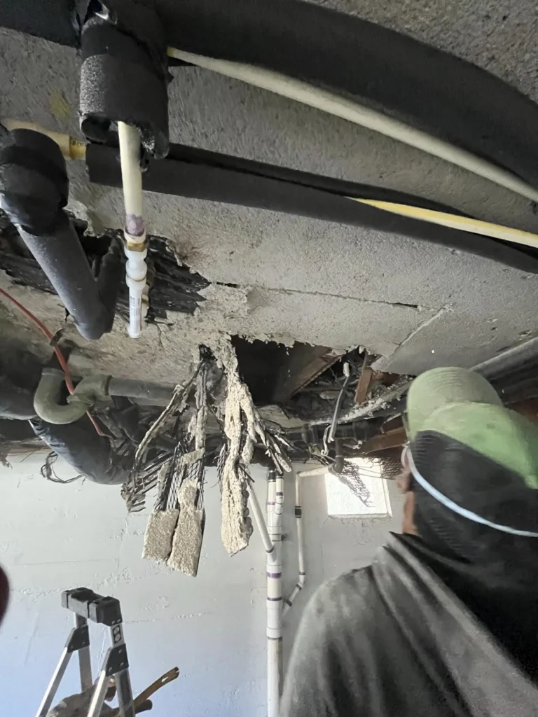 Basement ceiling with rotted wood joists and substantial fallen debris including plaster chunks, with black drain pipes, hanging utility lines, and daylight visible through basement windows below