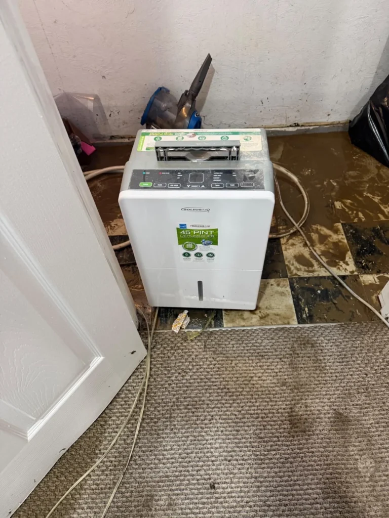 A Soleus Air 45-pint dehumidifier running in a corner of a flooded basement after water receded, showing severely damaged and discolored vinyl floor tiles, muddy residue, exposed dark subfloor, and drainage hoses
