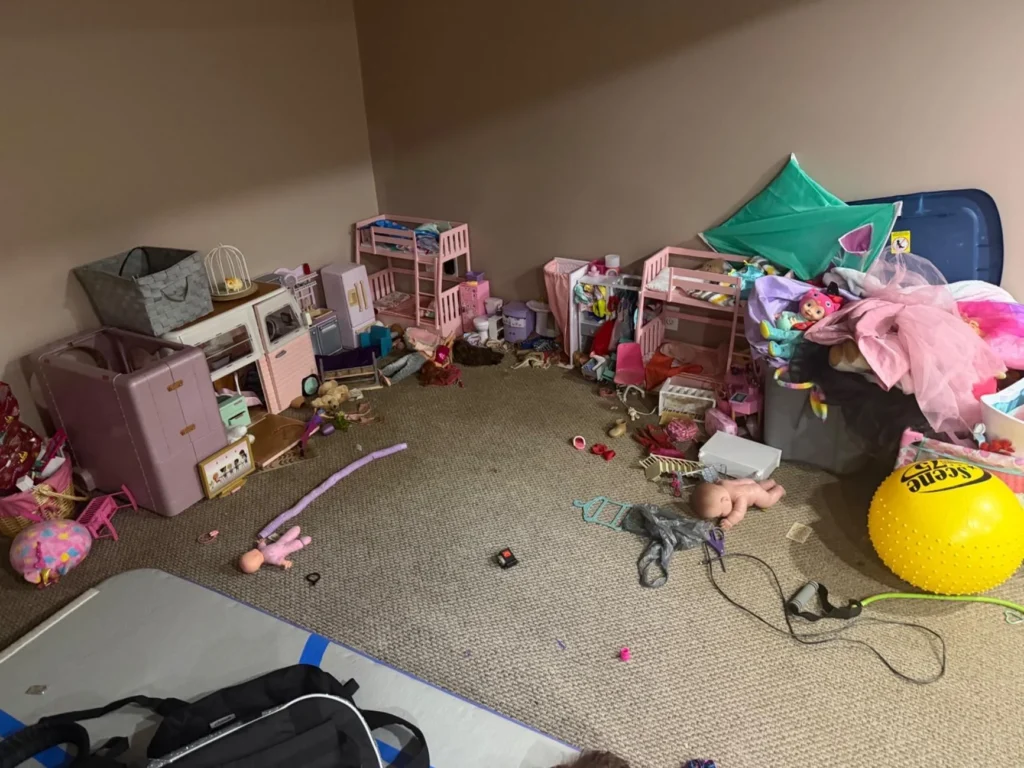 Basement playroom after flooding with the water receded, showing scattered children's toys including dollhouses, pink doll furniture, a baby doll, a yellow exercise ball, stuffed animals, and various debris on damp carpet
