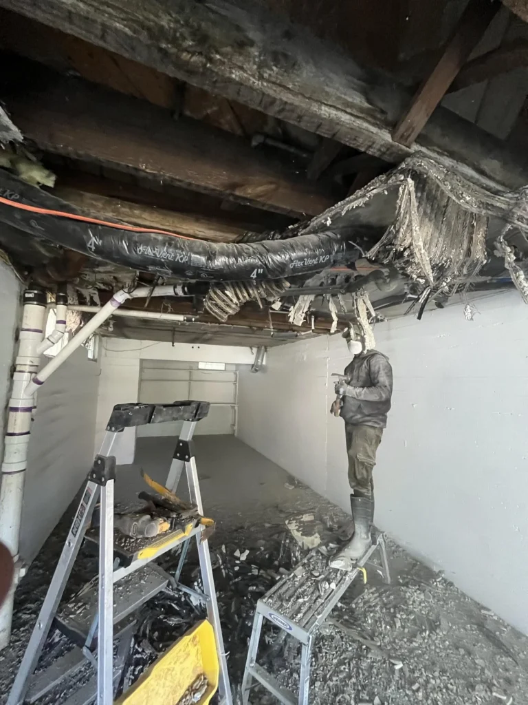 Remediation worker in mask and soiled clothing standing on a step stool while working on a severely damaged basement ceiling with a large black flexible duct, rotted wood joists, and dangling debris