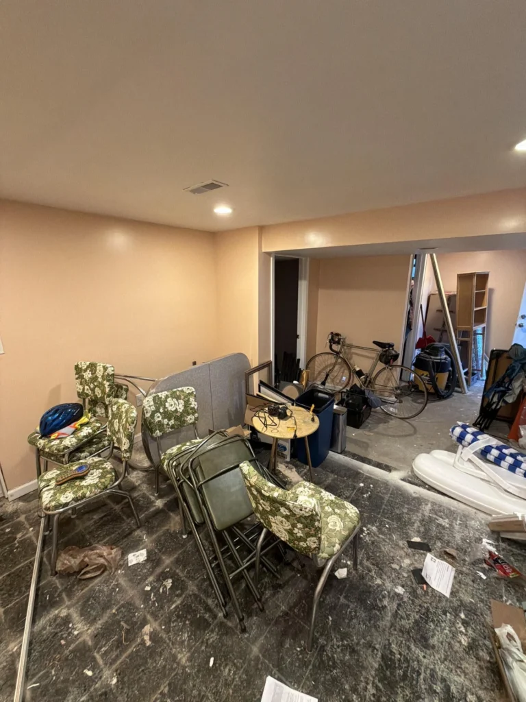 Basement room with water damage showing floral upholstered chrome chairs stacked and displaced, a bicycle, shelving unit, blue mattress and other contents moved to the center on a dirty damaged floor with peach-colored walls