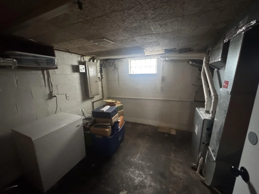 Basement utility room overview showing concrete block walls, drop ceiling tiles, electrical panel, HVAC ductwork with PVC pipes, a chest freezer, blue storage bin with boxes, and wet concrete floor